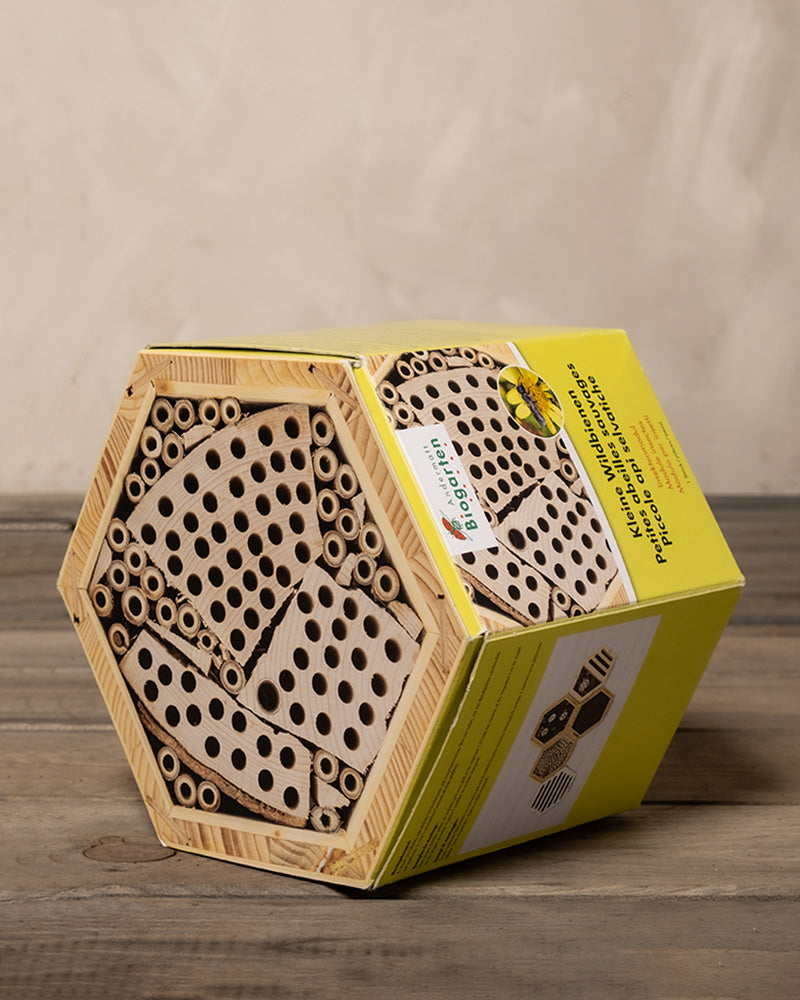 Sechseckiges Bienenhotel aus Holz mit gebohrten Löchern und Bambusrohren, das Solitärbienen anlocken soll. Das Bienenhotel steht auf einer Holzoberfläche und ist mit einer gelben Informationskarte mit Produktdetails und Informationen zum Artenschutz versehen.