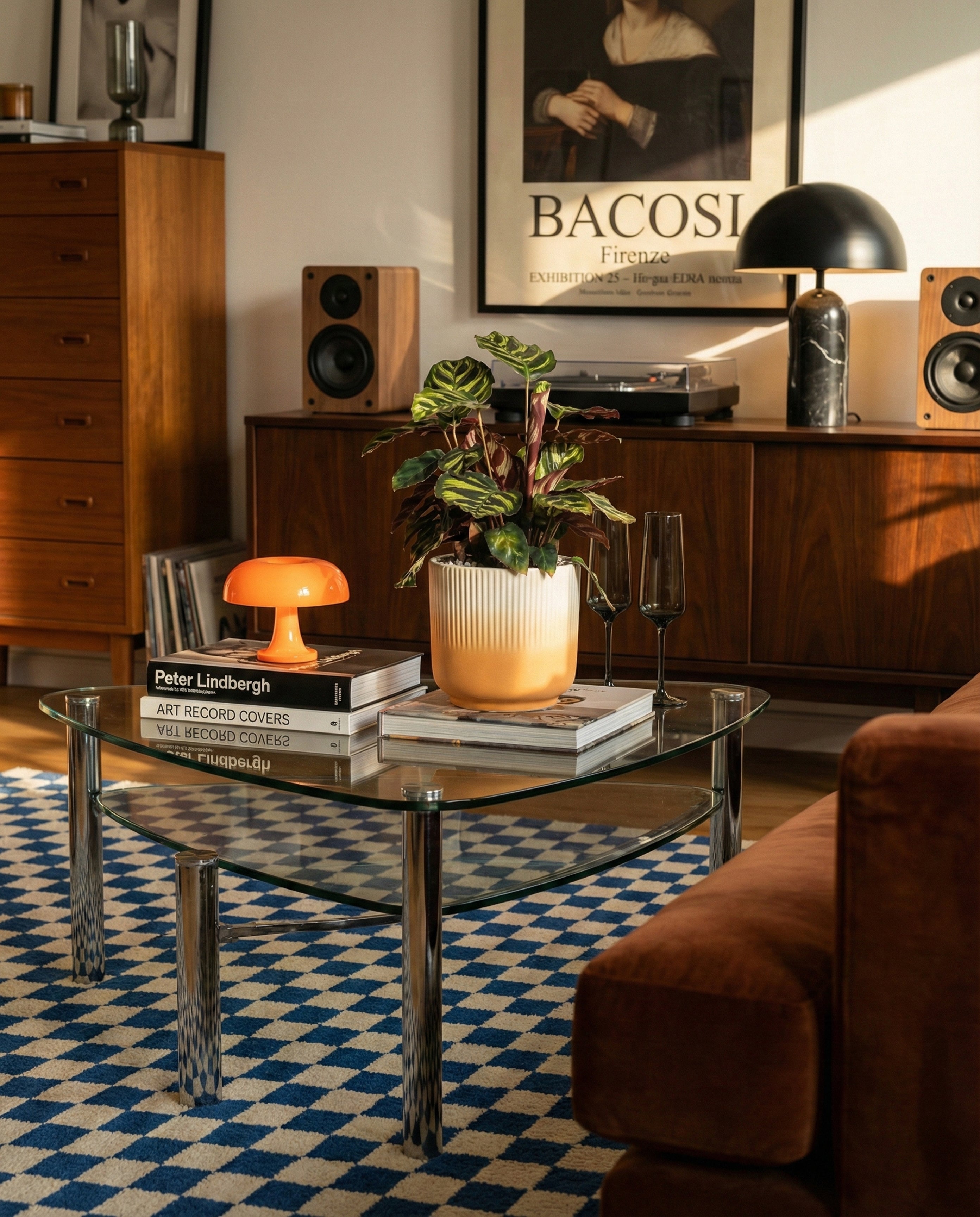 In einem stilvollen Wohnzimmer stehen ein gläserner Couchtisch mit Büchern, eine orangefarbene Lampe, die Kleine Calathea makoyana - feey und zwei Gläser. Ein blau-weißer Teppich, ein braunes Sofa und ein Holzschrank mit Lautsprechern und Kunstwerken vervollständigen den gemütlichen Raum.