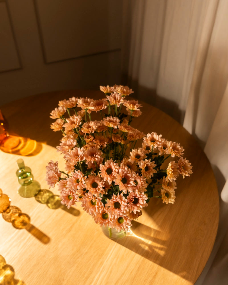 Chrysanthemen pfirsich (10 Stk.) in einer Glasvase stehen auf einem runden Holztisch im Sonnenlicht, schaffen eine blühende Atmosphäre mit Deko-Glasobjekten und transparenten Vorhängen im Hintergrund.