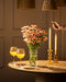 Eine Hand zündet eine Spitzkerze neben Chrysanthemen-Pfirsich (10 Stk.) und zwei Gläsern mit Orangengetränk auf einem runden Holztisch an und schafft eine blühende Atmosphäre unter warmem Licht und gewebten Lampenschirmen.