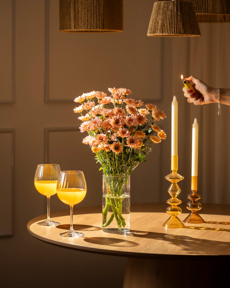 Eine Hand zündet eine Spitzkerze neben Chrysanthemen-Pfirsich (10 Stk.) und zwei Gläsern mit Orangengetränk auf einem runden Holztisch an und schafft eine blühende Atmosphäre unter warmem Licht und gewebten Lampenschirmen.