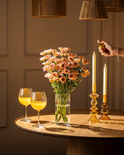 Eine Hand zündet eine Spitzkerze neben Chrysanthemen-Pfirsich (10 Stk.) und zwei Gläsern mit Orangengetränk auf einem runden Holztisch an und schafft eine blühende Atmosphäre unter warmem Licht und gewebten Lampenschirmen.