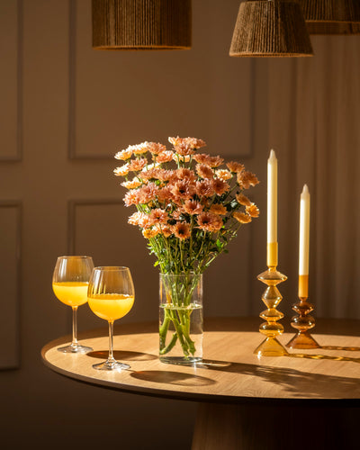 Ein runder Holztisch mit einer Vase mit Chrysanthemen-Pfirsich (10 Stk.), zwei Weingläsern mit Orangensaft und zwei brennenden gelben Kerzen in bernsteinfarbenen Haltern. Warmes Licht schafft eine blühende, unkomplizierte und einladende Atmosphäre.
