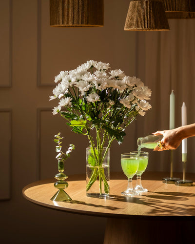 Ein Strauß Chrysanthemen weiß (5 Stk.) in einer schlanken Vase steht auf einem runden Holztisch. In der Nähe schenkt eine Hand ein grünes Getränk in elegante Gläser ein, begleitet von grünen Kerzen und einer kleinen Vase mit Grünzeug.