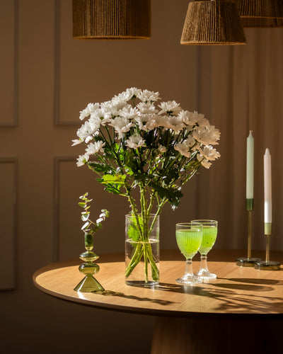 Ein Set Chrysanthemen weiß (5 Stk.) in einer schlanken Vase steht auf einem runden Holztisch mit zwei grünen Getränken, einer kleinen grünen Vase und zwei hohen Kerzen, die von oben warm beleuchtet werden.
