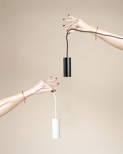 Zwei gepflegte Hände, die jeweils mit zarten Armbändern geschmückt sind, halten eine schwarze und eine weiße Pendelleuchte 'Corda' an der Kordel vor einem beigen Hintergrund - eine moderne Beleuchtung, die perfekt für Ihre Zimmerpflanzen ist.