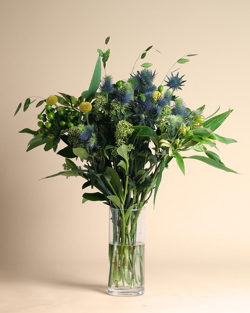 Die Vase Erfolgsgeheimnis zeigt ein klares Glasdesign, gefüllt mit grünen Blättern, gelben Bällen, blauen Disteln und strukturiertem Grün, das vor einem schlichten beigen Hintergrund positive Energie ausstrahlt.