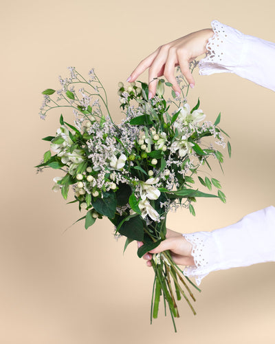 Eine Hand in einem weißen, mit Spitze besetzten Ärmel hält den Luftkuss-Strauß - grüne Blätter, weiße Blüten und kleine weiße Beeren - vor einem neutralen beigen Hintergrund.