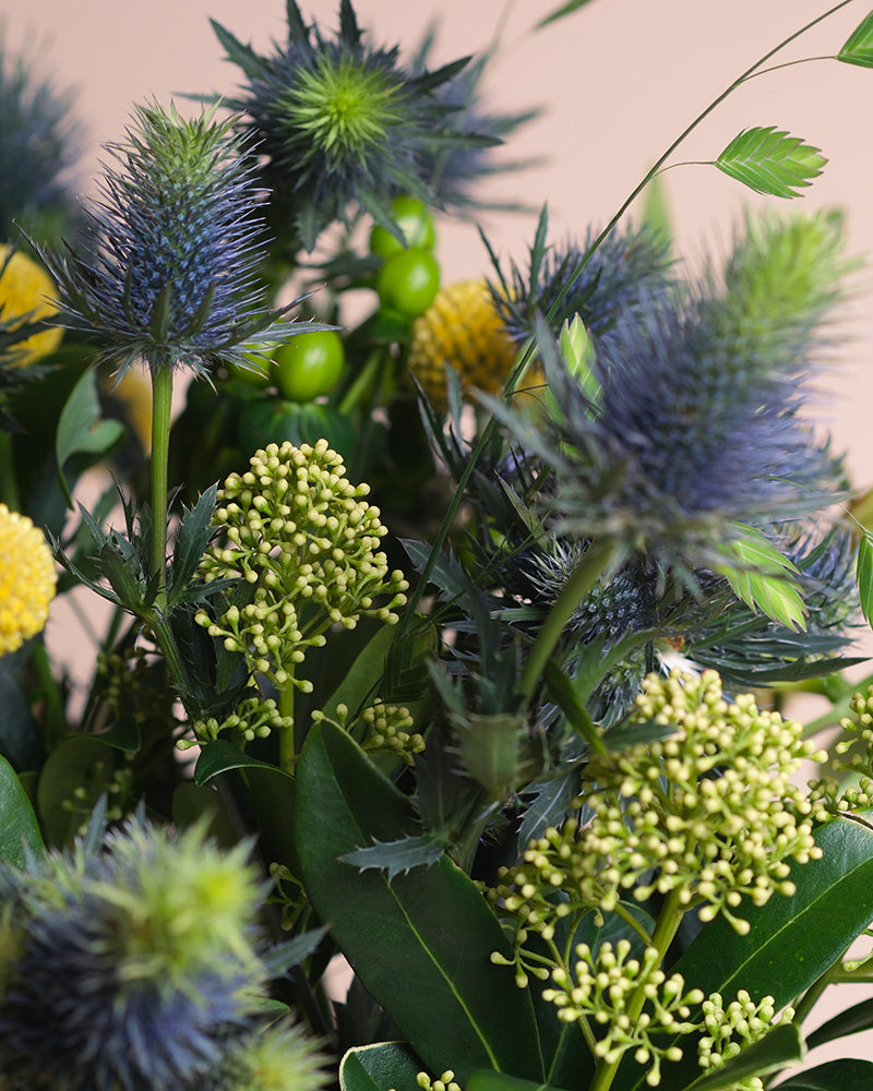 Eine Nahaufnahme des lebendigen Erfolgsgeheimnis-Straußes mit stacheligen blauen Disteln, runden gelben Blüten, grünen Beeren und üppigen Blättern, die positive Energie und Ruhe ausstrahlen.