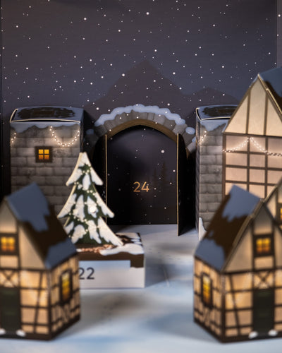 Eine festliche Szene mit dem Pflanzen Adventskalender mit bezaubernden Papierhäusern, einem schneebedeckten Weihnachtsbaum und einem Torbogen mit der Nummer „24“, der an einen Adventskalender erinnert. Im Hintergrund sorgen ein Sternenhimmel und schneebedeckte Berge für eine gemütliche, winterliche Atmosphäre.