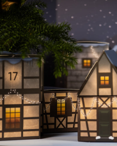 Eine festliche Szene zeigt den Pflanzen-Adventskalender mit beleuchteten Miniatur-Fachwerkhäusern, die mit Lichterketten geschmückt sind und sich vor einem Nachthimmel mit fallendem Schnee abheben. Das Haus im Vordergrund zeigt die Zahl 17 und hat eine grüne Tür, was an einen Adventskalender erinnert. Im Hintergrund ist ein Tannenzweig teilweise sichtbar.