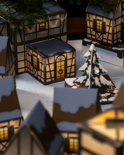 Eine gemütliche Miniatur-Winterdorfszene aus Papierhäusern mit schneebedeckten Dächern. Einige Häuser sind mit Lichterketten geschmückt und in der Mitte steht ein geschmückter Weihnachtsbaum. Aus den Fenstern leuchtet warmes gelbes Licht und sorgt für eine festliche Atmosphäre, die an den Pflanzen-Adventskalender erinnert.