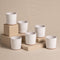 Ein Set aus sechs weißen Keramik-Baby-Topfsets „Sencillo“ (6 Stk.) mit braunem Boden, angeordnet auf beigen rechteckigen Blöcken vor einem beigen Hintergrund. Die Tassen sind gleichmäßig verteilt, einige auf höheren Blöcken und andere auf niedrigeren Flächen, wodurch eine ausgewogene Anordnung entsteht, die das schlichte Design hervorhebt.