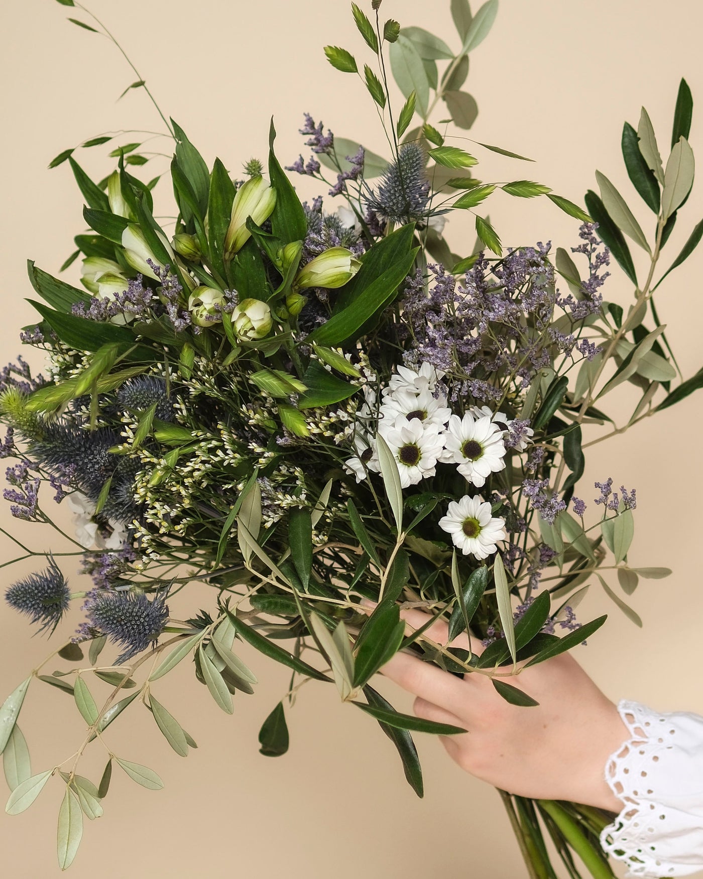 Eine Hand hält den Nebelmeer-Strauß - mit zarten Blüten wie weißen Gänseblümchen, lila Disteln, kleinen Lavendelblüten und grünen Blättern - vor einem hellbeigen Hintergrund.