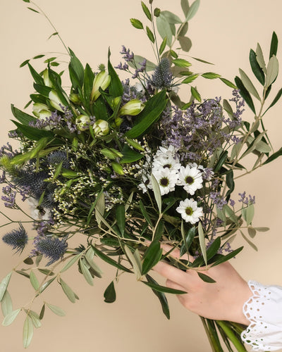 Eine Hand hält den Nebelmeer-Strauß - mit zarten Blüten wie weißen Gänseblümchen, lila Disteln, kleinen Lavendelblüten und grünen Blättern - vor einem hellbeigen Hintergrund.