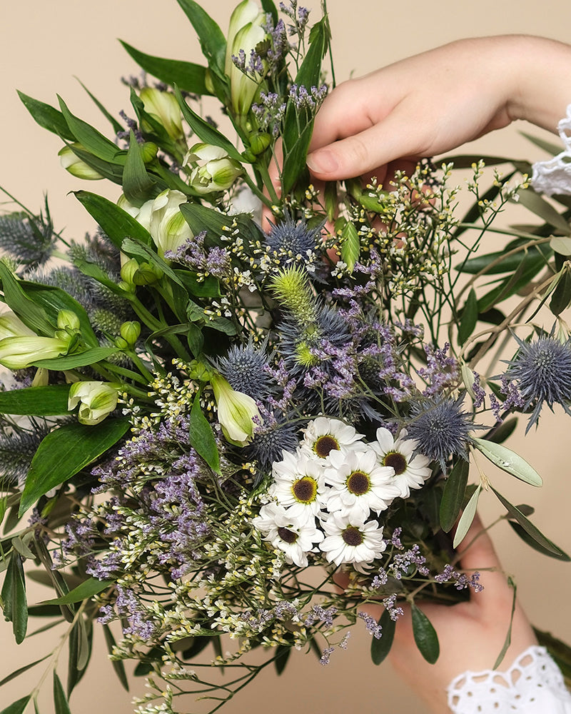 Eine Hand hält den Blumenstrauß "Nebelmeer" aus zarten Blüten mit kleinen weißen Gänseblümchen, violetten Disteln und grünen Blättern vor beigem Hintergrund.