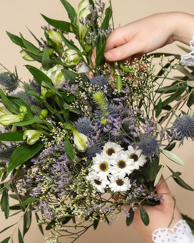 Eine Hand hält den Blumenstrauß "Nebelmeer" aus zarten Blüten mit kleinen weißen Gänseblümchen, violetten Disteln und grünen Blättern vor beigem Hintergrund.
