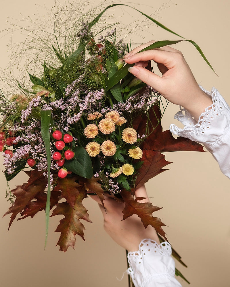 Eine Person in einer weißen Spitzenbluse arrangiert den Herbstfreude-Strauß - eine herbstliche Mischung aus gelben, roten und violetten Blumen mit großen braunen Blättern - vor einem beigen Hintergrund.