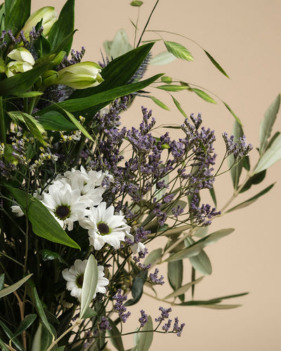 Ein zarter Blumenstrauß namens "Nebelmeer" aus weißen Gänseblümchen, violetten Blüten und grünen Blättern wirkt vor beigem Hintergrund wie frischer Morgentau am Morgen.