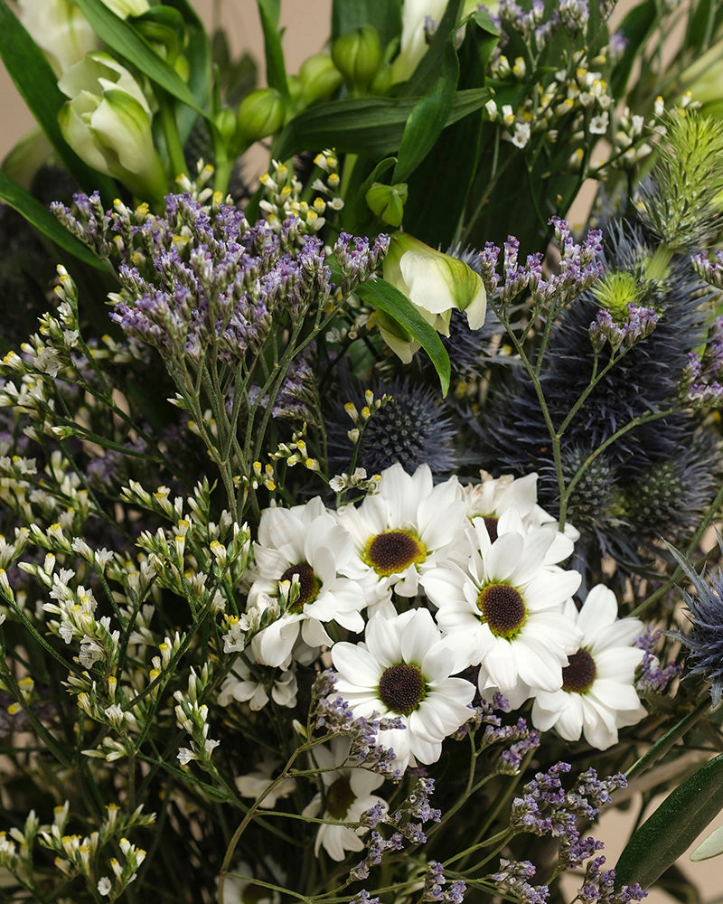 Der Blumenstrauß "Nebelmeer" vereint weiße Gänseblümchen mit gelben Zentren, zarte lila und weiße Blüten, grünes Laub und spitze blaue Disteln - wie von Morgentau geküsst.