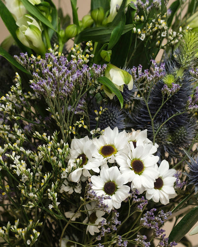 Der Blumenstrauß "Nebelmeer" vereint weiße Gänseblümchen mit gelben Zentren, zarte lila und weiße Blüten, grünes Laub und spitze blaue Disteln - wie von Morgentau geküsst.
