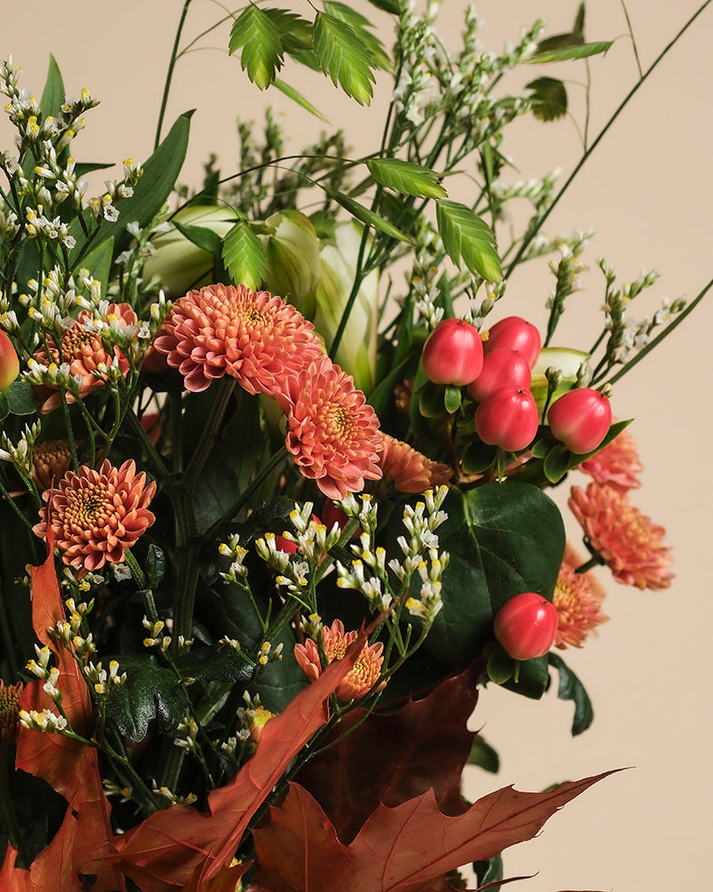 Herbstfreude zaubert herbstliche Stimmung: Mit orangefarbenen Chrysanthemen, pinken Hypericum-Beeren, weißen Blüten und grünem Laub schafft der Strauß ein fröhliches Farbspiel vor beigem Hintergrund.