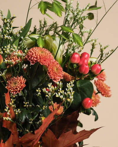 Herbstfreude zaubert herbstliche Stimmung: Mit orangefarbenen Chrysanthemen, pinken Hypericum-Beeren, weißen Blüten und grünem Laub schafft der Strauß ein fröhliches Farbspiel vor beigem Hintergrund.