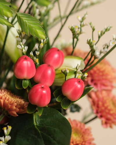 Das Herbstfreude Bouquet zeigt kleine, runde, pinke Beeren, grüne Blätter, weiße Knospen und orangefarbene Chrysanthemen in warmen Farbtönen - perfekt für den Herbst, vor sanft beigem Hintergrund arrangiert.