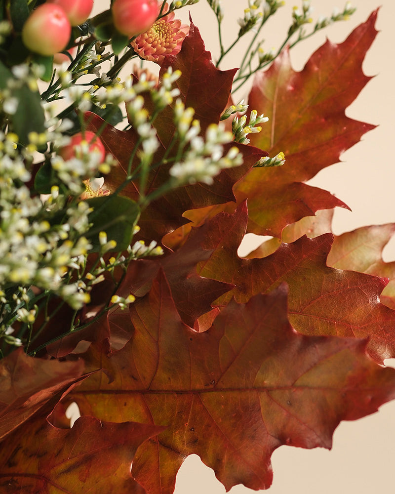 Nahaufnahme des Herbstfreude-Bouquets, das eine fröhliche Mischung aus rotbraunen Eichenblättern, kleinen weißen Blüten, grünen Stielen und Büscheln roter und rosa beerenartiger Akzente zeigt.
