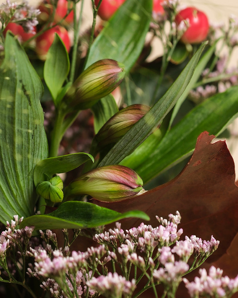Nahaufnahme des Straußes Herbstfreude in warmen Tönen: geschlossene rosa Lilien, üppige grüne Blätter, zarte kleine rosa Blüten und rote Beeren. Ein lebhaftes Farbenspiel unterstreicht die vielfältigen Texturen und reichen Farbtöne.
