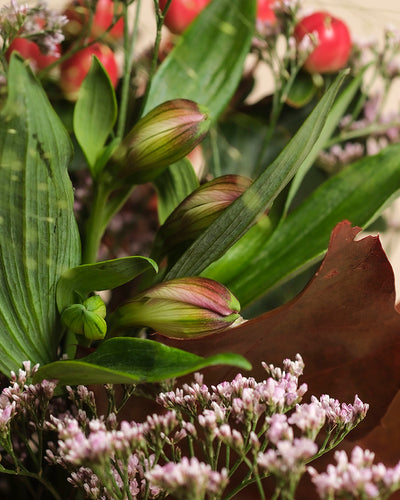 Nahaufnahme des Straußes Herbstfreude in warmen Tönen: geschlossene rosa Lilien, üppige grüne Blätter, zarte kleine rosa Blüten und rote Beeren. Ein lebhaftes Farbenspiel unterstreicht die vielfältigen Texturen und reichen Farbtöne.