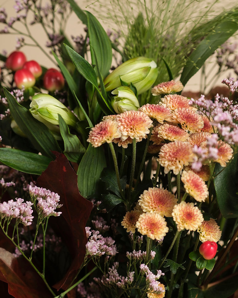 Herbstfreude: Ein fröhliches Farbspiel aus pfirsich- und pinkfarbenen Chrysanthemen, grünen Knospen, lila Blüten und roten Beeren, umrahmt von grünen Blättern.