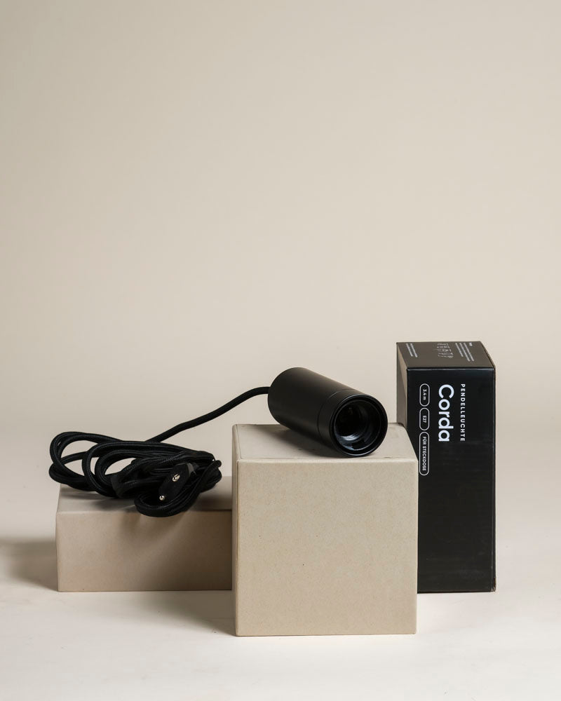 Eine schwarze Pendelleuchte 'Corda' mit einer Kordel ruht auf zwei beigen Kästen neben einer schwarzen "Coda"-Verpackung, alles vor einem hellen Hintergrund - perfekt als stilvolle Pflanzenlampe oder zum Kombinieren mit Zimmerpflanzen.