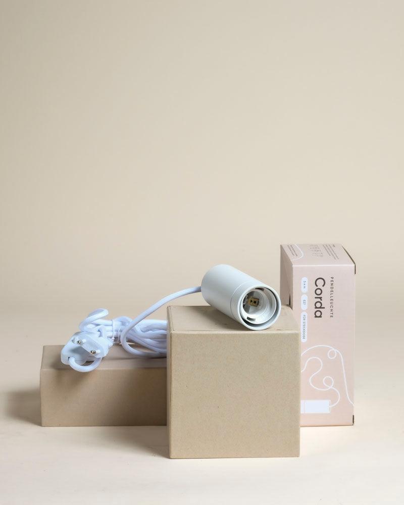 Die Pendelleuchte 'Corda' in Weiß, mit Kordel und Steckdose, wird auf beigen Kartons mit ihrer Verpackung im Hintergrund präsentiert - ideal als stilvolle Pflanzenlampe für Ihre Zimmerpflanzen.