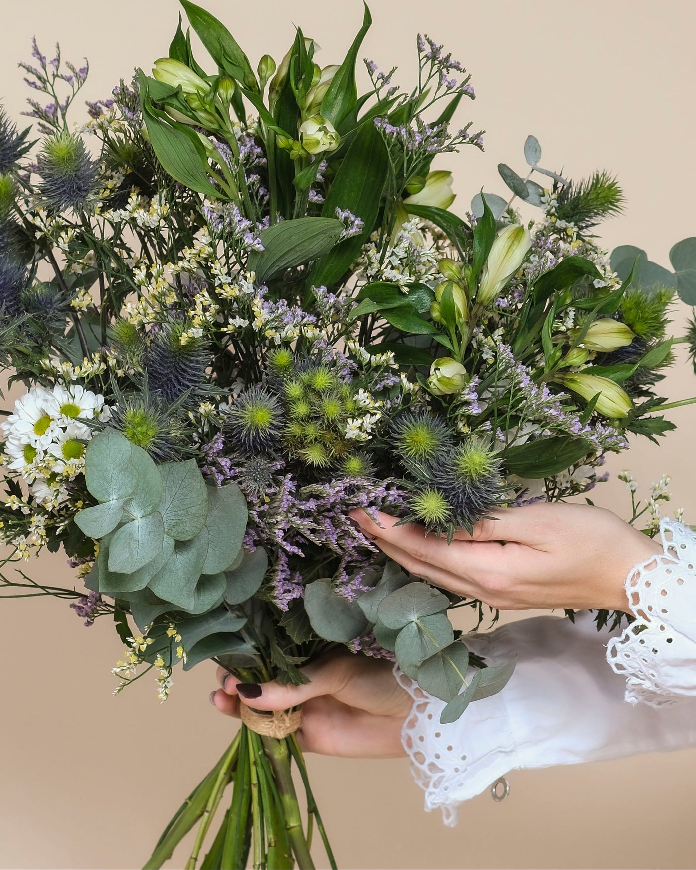 Eine Person in einem weißen Hemd mit Spitzenärmeln hält den Nebelmeer-Strauß - mit grünen Blättern, violetten Disteln, Morgentau, zarten Blüten und weißen Gänseblümchen - vor einem beigen Hintergrund.