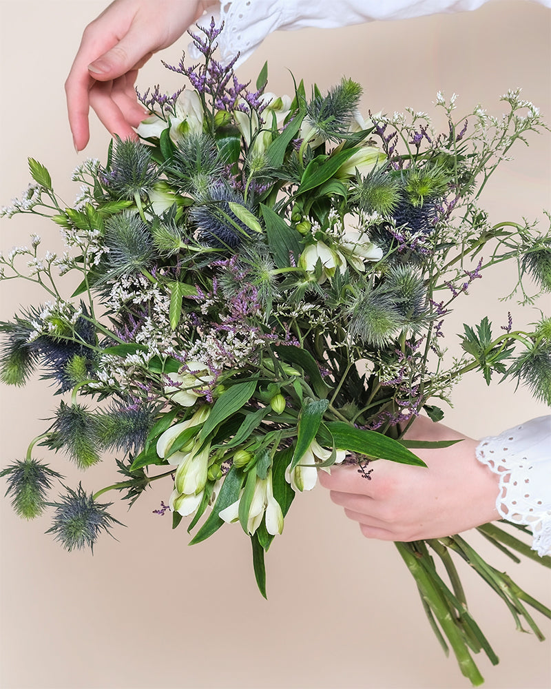 Eine Person in einem weißen Hemd mit Ösenmanschetten hält den "Wanderlust"-Blumenstrauß - grüne Blätter, weiße Blütenblätter und stachelige blaue distelähnliche Blumen - in der Hand, um die Natur vor einem hellbeigen Hintergrund in Szene zu setzen.