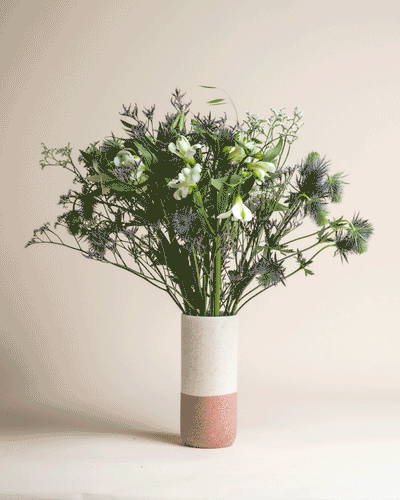 Die Keramikvase "Wanderlust" mit weißem und rosafarbenem Blockdesign enthält grüne und weiße Blumen, üppiges Grün und blaue Disteln, die an die Frische der Bergluft vor einem schlichten beigen Hintergrund erinnern.
