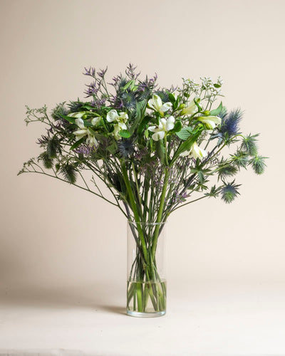 Der Strauß Wanderlust besteht aus grünen, weißen und violetten Blumen mit langen Stielen und stacheligen Blättern in einer klaren Glasvase, die an die reine Natur vor einem schlichten, hellen Hintergrund erinnert.