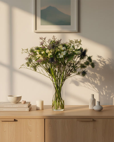 In einer klaren Glasvase steht der grün-weiße Wanderlust-Strauß und bringt Bergluft-Stimmung in einen hellen Holzschrank. In der Nähe liegen Bücher, Kerzen und kleine Vasen, während das Sonnenlicht sanfte Schatten unter die gerahmte Wandkunst wirft.