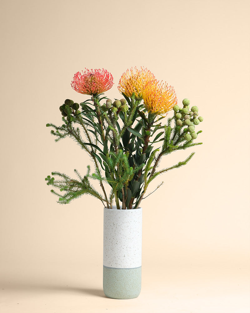 Die Vase Couture zeigt einen stilvollen Strauß Protea-Blüten mit orangefarbenen Blütenblättern und grünem Laub, eingebettet in ein modernes weißes und hellblau gesprenkeltes Design vor einem schlichten beigen Hintergrund.