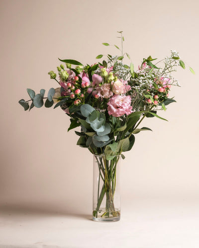 In einer klaren Glasvase steht der Ballerina-Strauß mit rosa Blumen, Eukalyptusblättern und Grünzeug vor einem schlichten beigen Hintergrund.