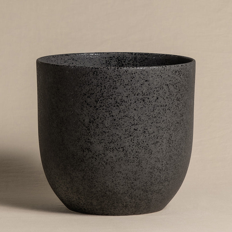 Eine schwarze, runde, gesprenkelte Keramikvase mit einer weiten Öffnung oben, abgebildet vor einem neutralen beigen Hintergrund. Der fey Keramik-Topf (Direito | 22 cm ⌀) besticht durch ein minimalistisches, modernes Design mit einer glatten Textur.