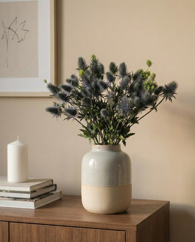 Eine Edeldistel-Keramikvase mit blauen Stachelblüten steht auf einem Holzschrank neben einer weißen Kerze und gestapelten Büchern, vor einer neutralen Wand mit gerahmter minimalistischer Kunst.