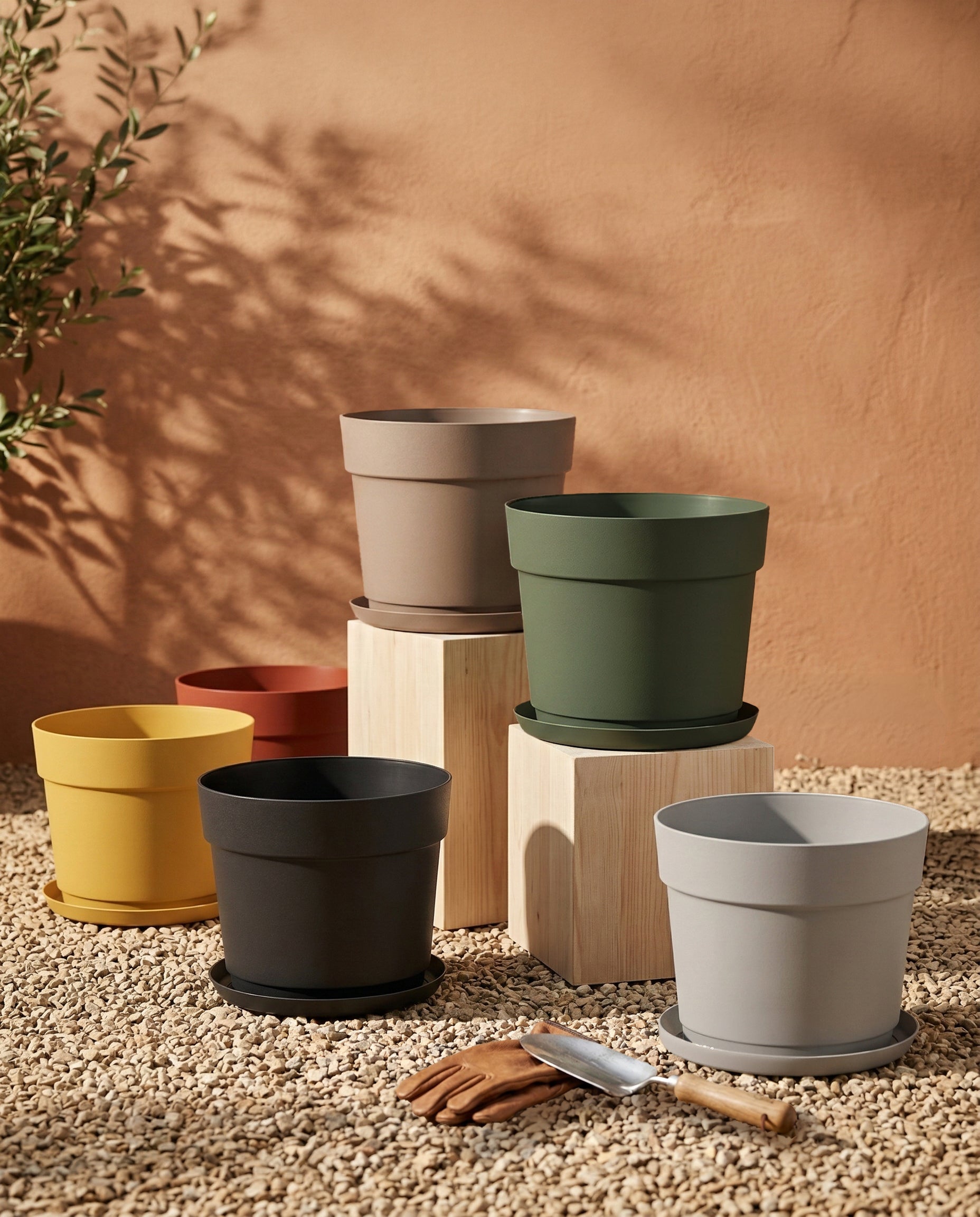 Sechs farbenfrohe Kunststoff Pflanzkübel (Bloom | 29 cm Ø) in grau, grün, schwarz, gelb und rot stehen auf hellen Holzklötzen und Kies. Hergestellt aus recyceltem Kunststoff und UV-beständig, passen sie in jeden Außenbereich. Im Hintergrund ist ein Olivenbaum zu sehen.