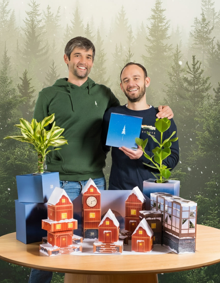 Zwei lächelnde Männer stehen hinter einem Tisch mit getopften Zimmerpflanzen und Miniaturhäusern. Einer hält eine blaue Pflanzen Adventskalender | NIKIN-Special (Dschungel-Version) Box. Im Hintergrund ist eine verschneite, immergrüne Waldszene zu sehen.
