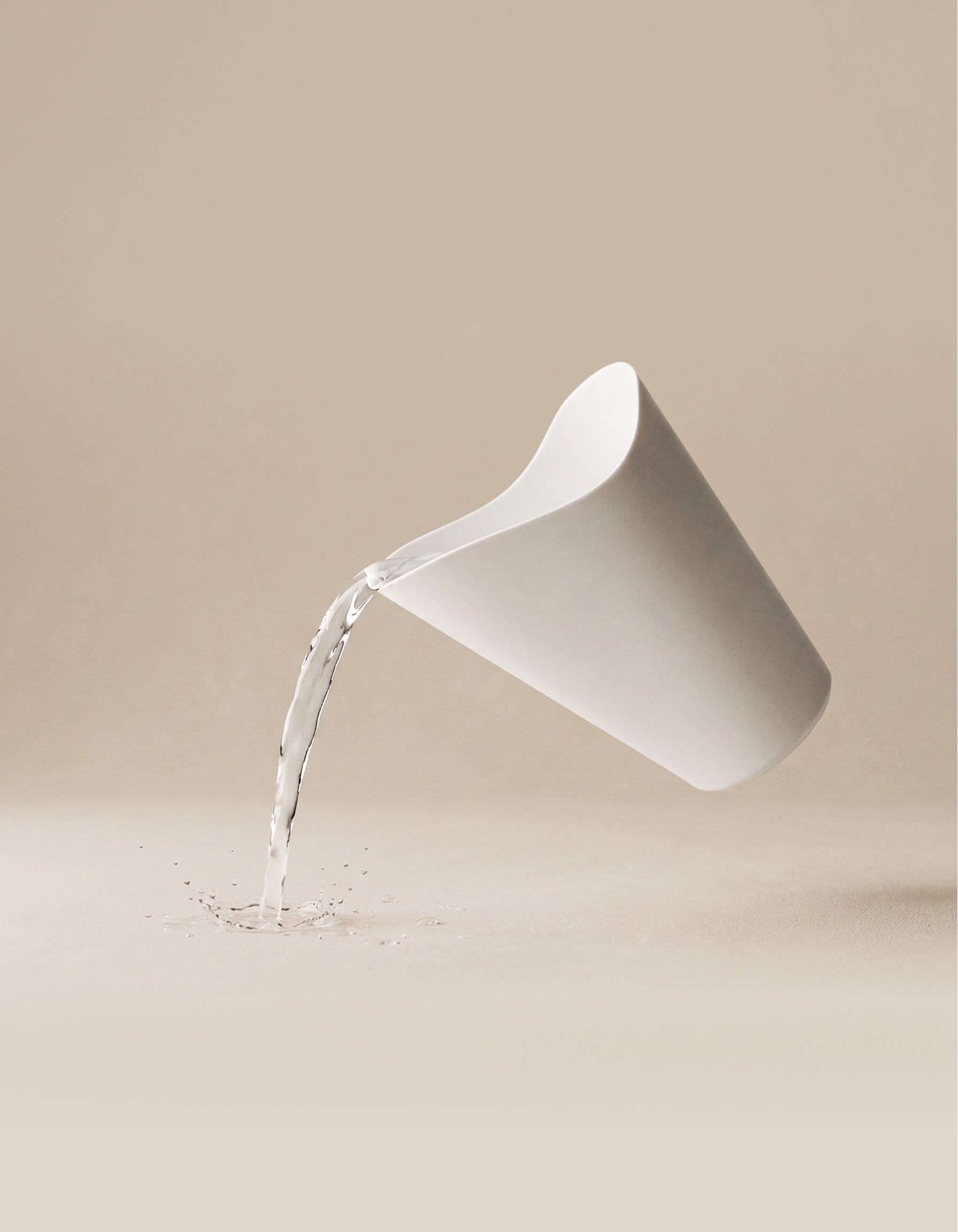 Ein weißer Giessbecher für Babypflanzen neigt sich in der Luft und gießt mit einem sanften Spritzer Wasser auf eine glatte, beigefarbene Oberfläche; die Kulisse ist neutral und minimalistisch.