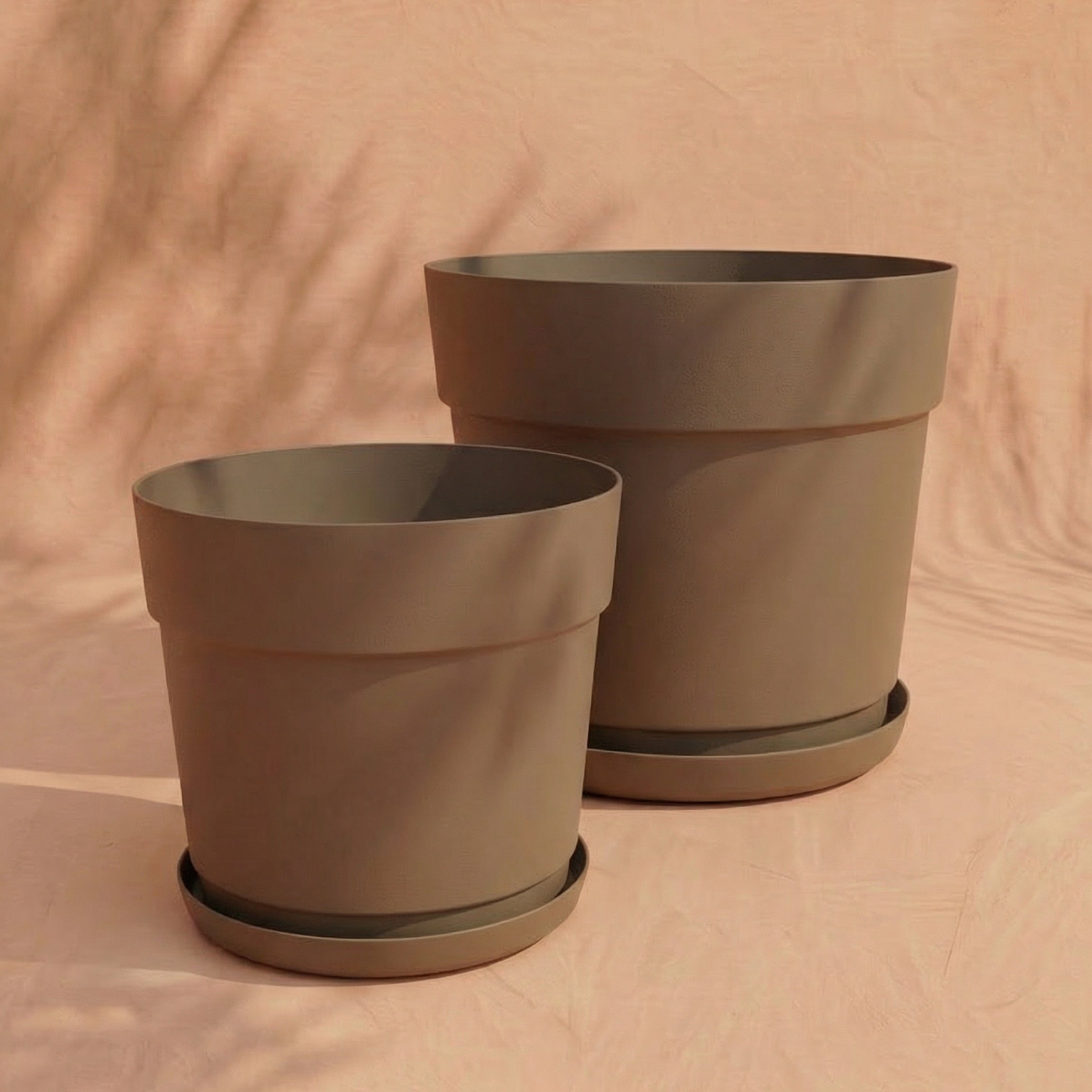 Zwei leere, hellbraune Pflanzkübel-Sets 'Bloom' (38, 29) aus recyceltem Kunststoff mit passenden Untertassen stehen nebeneinander auf einer beigen Fläche und werfen weiche, natürliche Schatten in den Hintergrund.