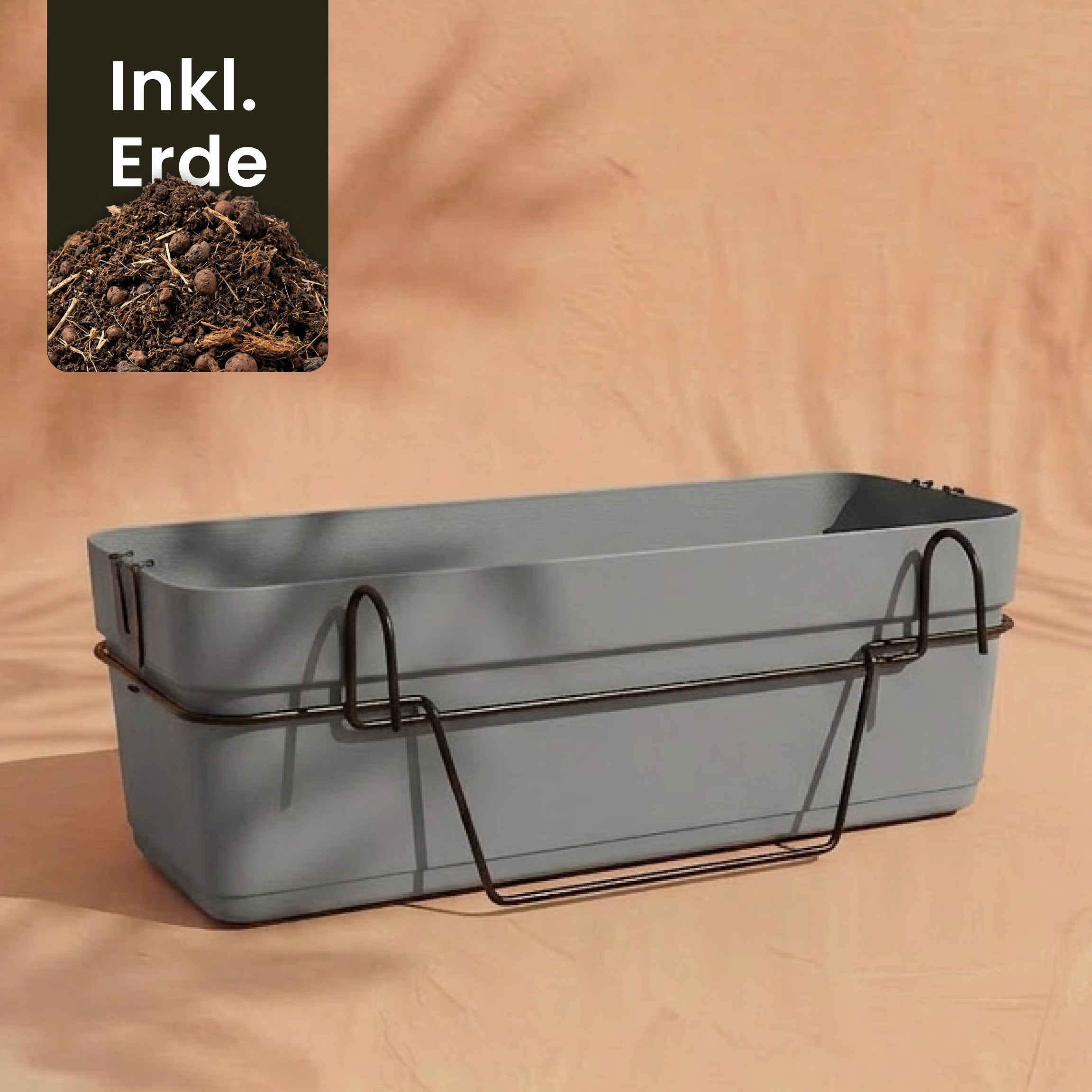 Ein rechteckiger grauer Pflanzkasten für Geländer (Blossom | 60 cm) mit Wasserreservoir und schwarzem Metallhalter steht auf einem pfirsichfarbenen Hintergrund. Ein Einsatz zeigt Erde mit der Aufschrift 
