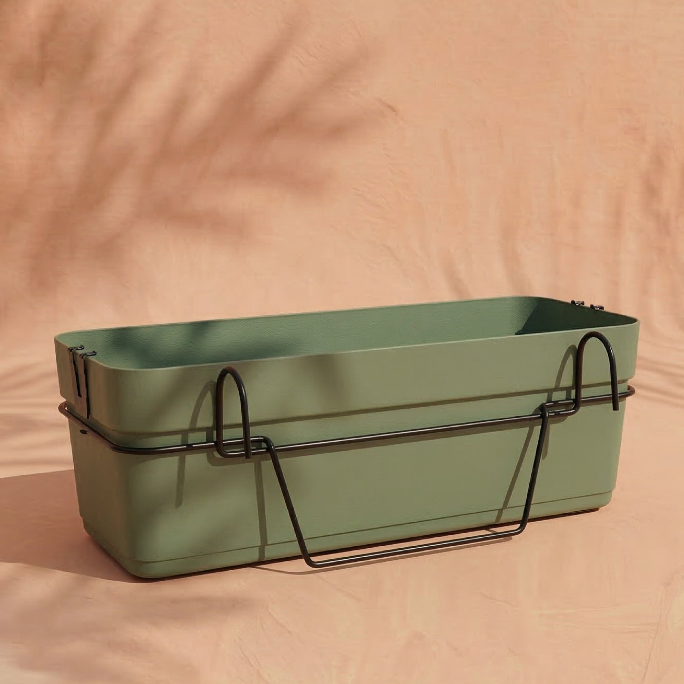 Ein rechteckiger grüner Pflanzkasten für Geländer (Blossom | 60 cm) mit einem schwarzen Metallhalter ruht auf einer pfirsichfarbenen Oberfläche, ideal für Ihren Balkon. Weiche Schatten im Hintergrund unterstreichen sein stilvolles Aussehen.