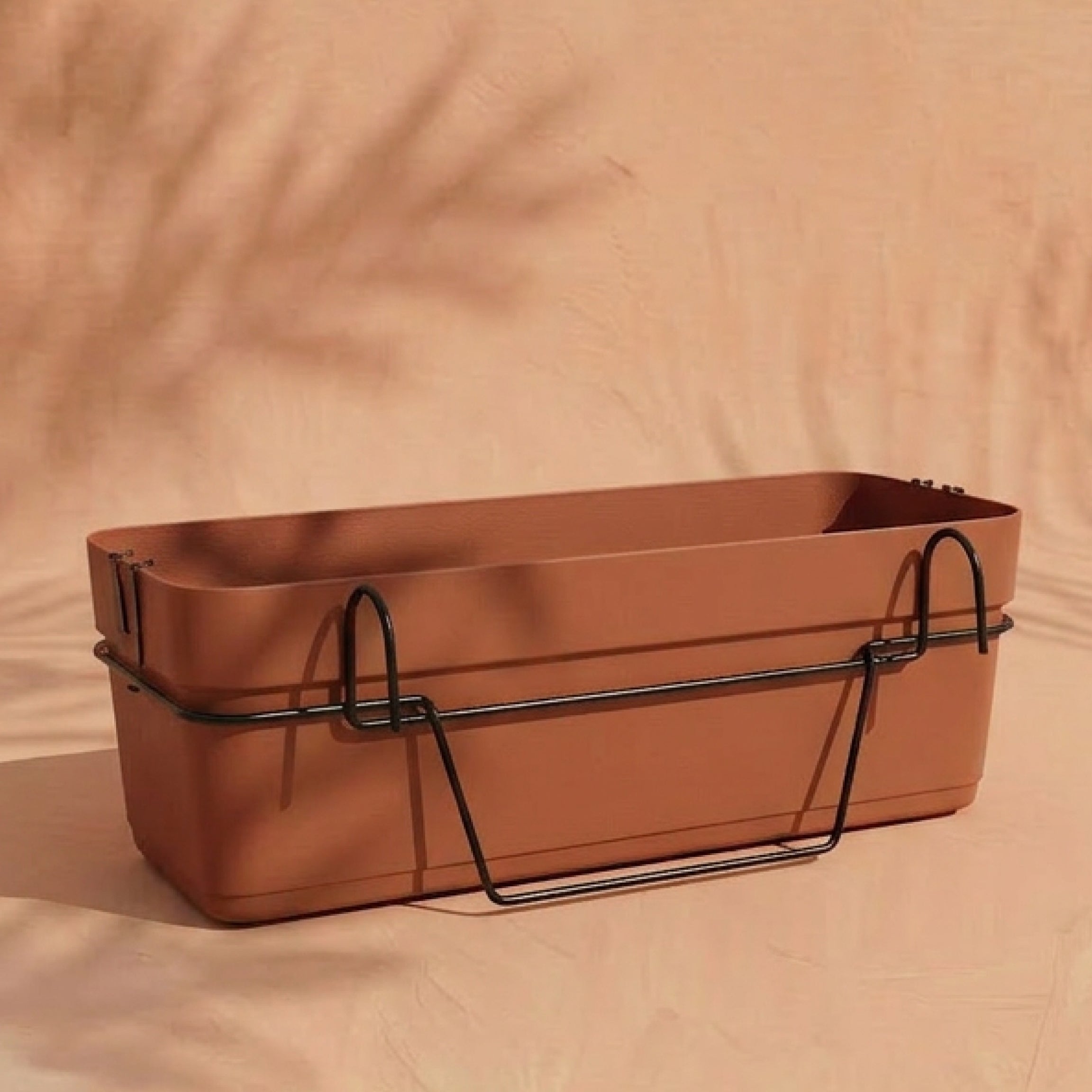 Der Pflanzkasten für Geländer (Blossom | 60 cm) in Terrakotta mit Wasserreservoir und schwarzer Metall-Halterung steht auf einer hellen Pfirsichoberfläche, sanfte Blattschatten im Hintergrund - ideal für den Balkon.